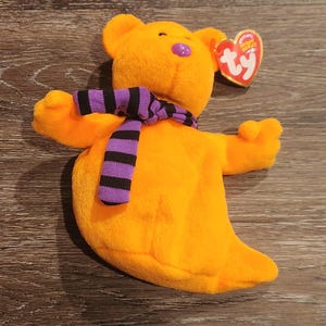 May include: A bright orange plush bear toy, featuring a purple nose and a black and purple striped scarf. The bear has a Ty heart-shaped tag attached. The toy is made of soft, plush material.