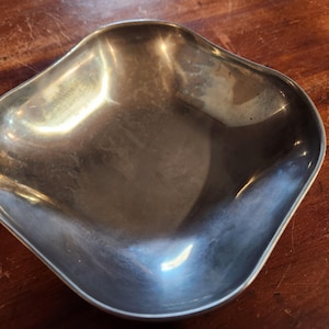 May include: A silver-colored, four-lobed bowl with a smooth, reflective surface. The bowl is empty and sits on a dark wooden surface. The bowl's shape is reminiscent of a flower or a stylized quatrefoil.