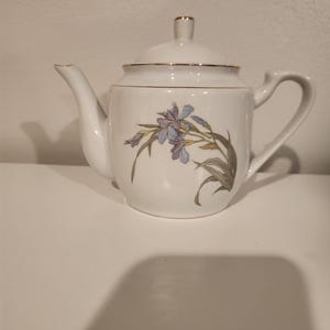 May include: White ceramic teapot with a lid and handle. The teapot features a floral design with purple and blue flowers and green leaves. Gold trim accents the lid and rim of the teapot.
