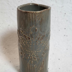 May include: A gray ceramic vase with a textured, embossed design. The vase has a cylindrical shape and a slightly flared rim.