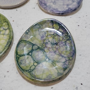 May include: A small, oval-shaped ceramic dish with a green, purple, and white marbled glaze. The glaze has a bubbled effect, creating a unique and textured surface.