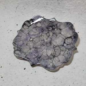 May include: A decorative ceramic dish with a scalloped edge and a white, gray, and purple marbled design. The dish has a glossy finish and features a pattern of black lines and bubbles.