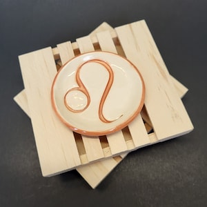 May include: A white ceramic dish with a brown outline and a Leo zodiac symbol in the center. The dish is sitting on a wooden stand.