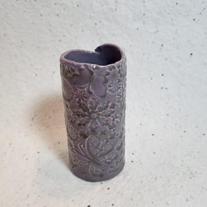 May include: A purple ceramic vase with a floral and butterfly design. The vase is shaped like a heart at the top and has a cylindrical body.