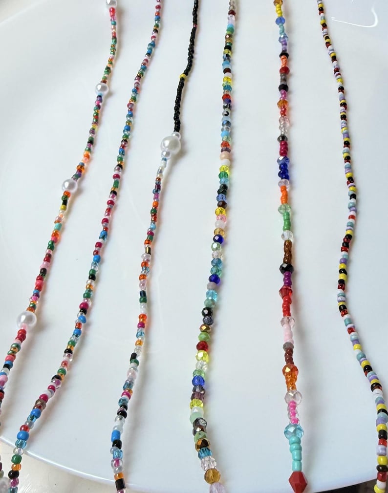 May include: A selection of beaded necklaces and eyeglass chains in various colours. The strands are made up of small, colourful beads, with larger pearl-like beads interspersed. The necklaces are displayed on a white surface.