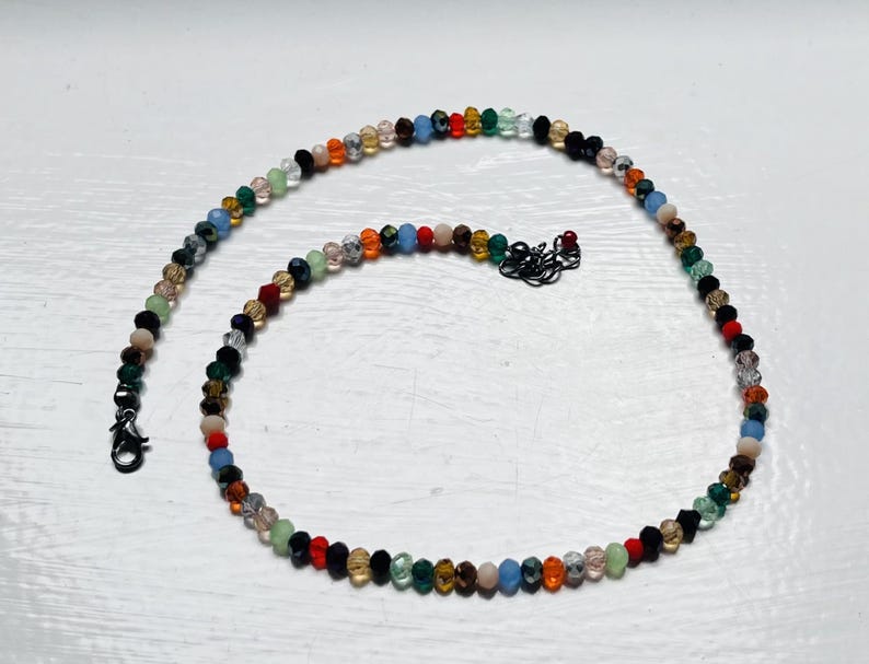 May include: A necklace made of small, colourful beads. The beads are in shades of red, orange, yellow, green, blue, and black. The necklace has a black clasp and measures approximately 40 cm in length.