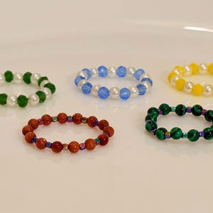 May include: Five beaded rings in different colours: green and white, blue and white, yellow and white, red and orange, and green and black. The rings are made of small beads strung together in a circular shape.