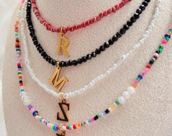 May include: Four beaded necklaces with gold initial pendants. The top necklace is red, followed by black, white, and a colourful beaded necklace. The gold pendants feature the letters R, M, S, and D.