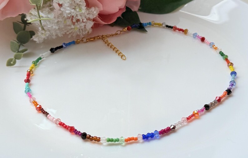 May include: A multi-coloured beaded necklace with a gold-coloured clasp. The necklace features small, round beads in red, orange, yellow, green, blue, and purple. The beads are strung together to create a delicate and eye-catching accessory.
