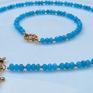 Könnte beinhalten: Ein Schmuckset bestehend aus einer Halskette und einem Armband aus kleinen, facettierten, leuchtend blauen Perlen mit goldfarbenen Zwischenelementen. Die Halskette hat einen goldfarbenen Knebelverschluss und einen Schildkrötenanhänger mit blau-weißem Muster.
