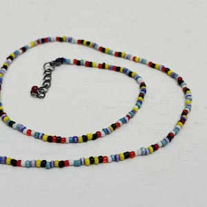 May include: A multi-coloured beaded necklace featuring small, round beads in red, yellow, blue, black, and white. The necklace has a black clasp and an adjustable chain. The beads are strung together to create a vibrant accessory, perfect for adding a pop of colour.