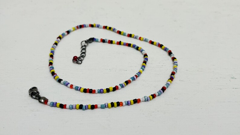 May include: A multi-coloured beaded necklace featuring small, round beads in red, yellow, blue, black, and white. The necklace has a black clasp and an adjustable chain. The beads are strung together to create a vibrant accessory, perfect for adding a pop of colour.