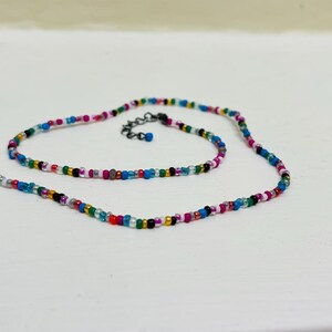 May include: A colourful beaded necklace featuring small, round beads in pink, blue, green, yellow, and black. The necklace has a silver-coloured clasp and a small extension chain. The beads are strung together to create a vibrant accessory.