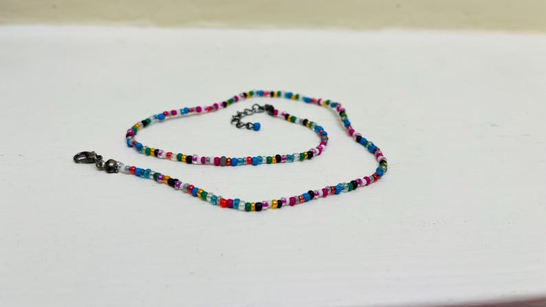 May include: A colourful beaded necklace featuring small, round beads in pink, blue, green, yellow, and black. The necklace has a silver-coloured clasp and a small extension chain. The beads are strung together to create a vibrant accessory.
