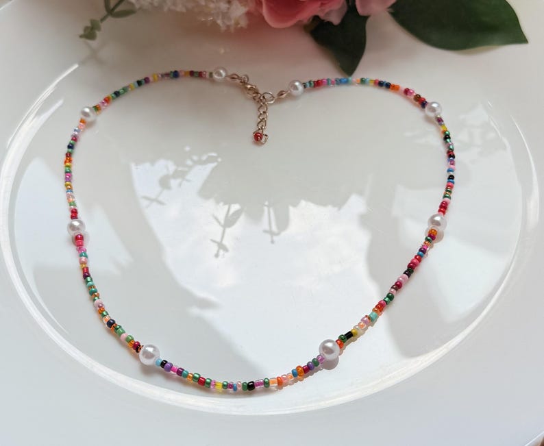 May include: A vibrant beaded necklace featuring a spectrum of small beads and white pearls. The necklace is presented on a white plate, with a floral arrangement visible in the background.