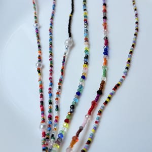 May include: Close-up of several beaded necklaces in various colours. The necklaces feature small, colourful beads interspersed with larger, pearl-like beads. The beads are strung on thin cords, creating a delicate and intricate design.