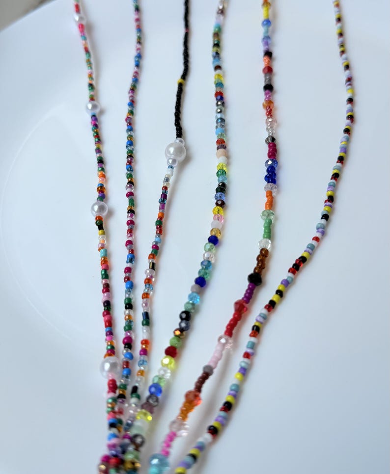 May include: Close-up of several beaded necklaces in various colours. The necklaces feature small, colourful beads interspersed with larger, pearl-like beads. The beads are strung on thin cords, creating a delicate and intricate design.