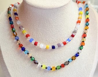 May include: Two colourful beaded necklaces displayed on a white bust. The necklaces feature a variety of square beads in red, orange, yellow, green, blue, and purple. The beads are strung together with black spacers.