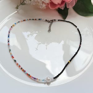 May include: A colourful beaded necklace featuring a mix of small beads and a section of black beads. A large white pearl is centered on the necklace. The necklace is on a white plate with a pink flower in the background.