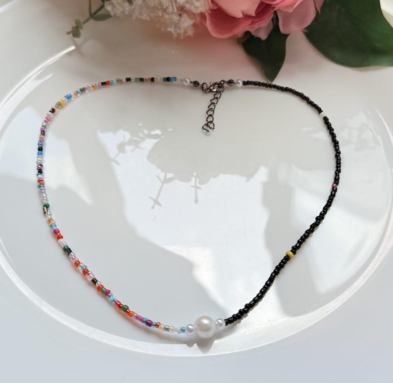 May include: A colourful beaded necklace featuring a mix of small beads and a section of black beads. A large white pearl is centered on the necklace. The necklace is on a white plate with a pink flower in the background.