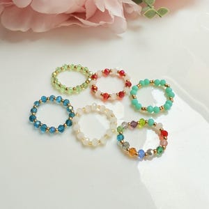 May include: A collection of beaded rings in various colors, including green, red, turquoise, blue, white, and rainbow. Each ring features small, colorful beads strung together with gold-colored accents. The rings are displayed on a white surface.