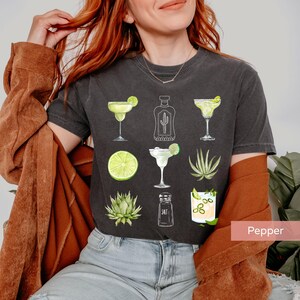 May include: A dark gray t-shirt with a graphic design of a margarita, lime, agave plant, tequila bottle, salt shaker, and pepper. The design is in a watercolor style.