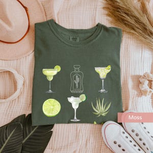 May include: A green t-shirt with a white outline of a bottle of tequila, two margarita glasses, a lime, and an agave plant. The text "Moss" is visible in the lower right corner of the image.