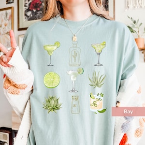 May include: A light blue t-shirt with a graphic design of various margarita-themed illustrations, including a bottle of tequila, limes, salt, and agave plants.