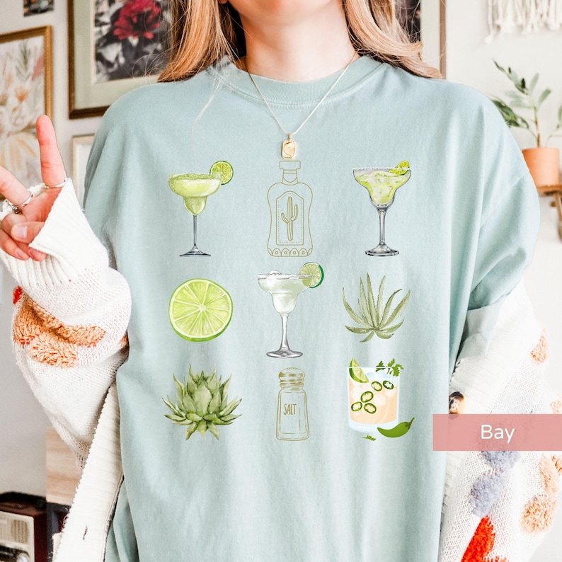 May include: A light blue t-shirt with a graphic design of various margarita-themed illustrations, including a bottle of tequila, limes, salt, and agave plants.