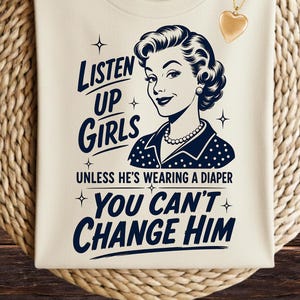 May include: A cream-coloured t-shirt featuring a dark blue graphic of a woman with a pearl necklace and the text: LISTEN UP GIRLS UNLESS HE'S WEARING A DIAPER YOU CAN'T CHANGE HIM. A gold heart-shaped pendant necklace is placed on the upper right side.