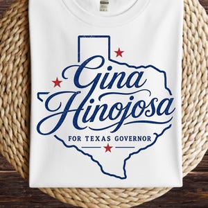 May include: A white t-shirt featuring a blue outline of the state of Texas with the text Gina Hinojosa for Texas Governor in blue script and block lettering, accented by three small red stars positioned around the state map graphic.