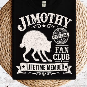 May include: A black t-shirt featuring a white graphic of a raccoon and the text JIMOTHY OFFICIAL SHORT SPINED RACCOON FAN CLUB LIFETIME MEMBER. The design includes a circular badge, stars, and decorative flourishes surrounding the central animal illustration.