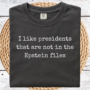 May include: Dark gray t-shirt with the text "I like presidents that are not in the Epstein files" in white. The shirt is folded and rests on a woven, circular placemat, set against a white wooden background.
