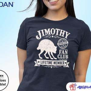 May include: A navy blue short-sleeved t-shirt featuring a white graphic of a raccoon. The text reads JIMOTHY OFFICIAL RACCOON FAN CLUB LIFETIME MEMBER. The design includes a circular badge and a banner element with a distressed, weathered texture.
