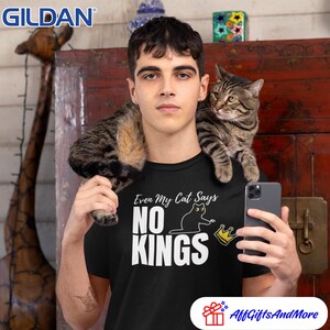 Puede incluir: Una camiseta negra de manga corta con el texto en blanco Even My Cat Says NO KINGS, acompa&ntilde;ado de un gr&aacute;fico de un gato negro y una corona amarilla. El logotipo de la marca Gildan es visible en la esquina superior izquierda de la imagen.