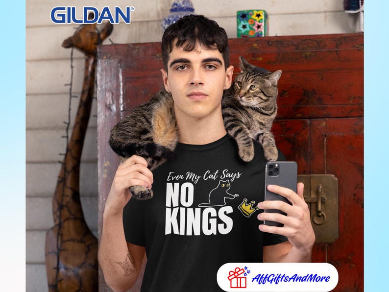 Puede incluir: Una camiseta negra de manga corta con el texto en blanco Even My Cat Says NO KINGS, acompa&ntilde;ado de un gr&aacute;fico de un gato negro y una corona amarilla. El logotipo de la marca Gildan es visible en la esquina superior izquierda de la imagen.