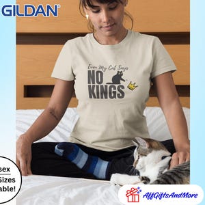 Puede incluir: Una camiseta beige de manga corta con un estampado gr&aacute;fico de la silueta de un gato negro y una peque&ntilde;a corona amarilla. El texto dice Even My Cat Says NO KINGS en una fuente gris oscuro. La camiseta es de la marca Gildan con cuello redondo.