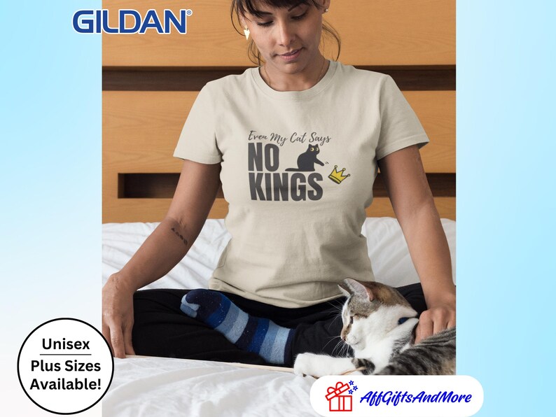 Puede incluir: Una camiseta beige de manga corta con un estampado gr&aacute;fico de la silueta de un gato negro y una peque&ntilde;a corona amarilla. El texto dice Even My Cat Says NO KINGS en una fuente gris oscuro. La camiseta es de la marca Gildan con cuello redondo.
