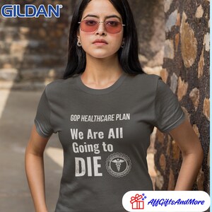 May include: Dark gray t-shirt with the text "GOP HEALTHCARE PLAN We Are All Going to DIE" in white. The shirt features a medical symbol and the Gildan logo. The model is wearing pink-tinted sunglasses.
