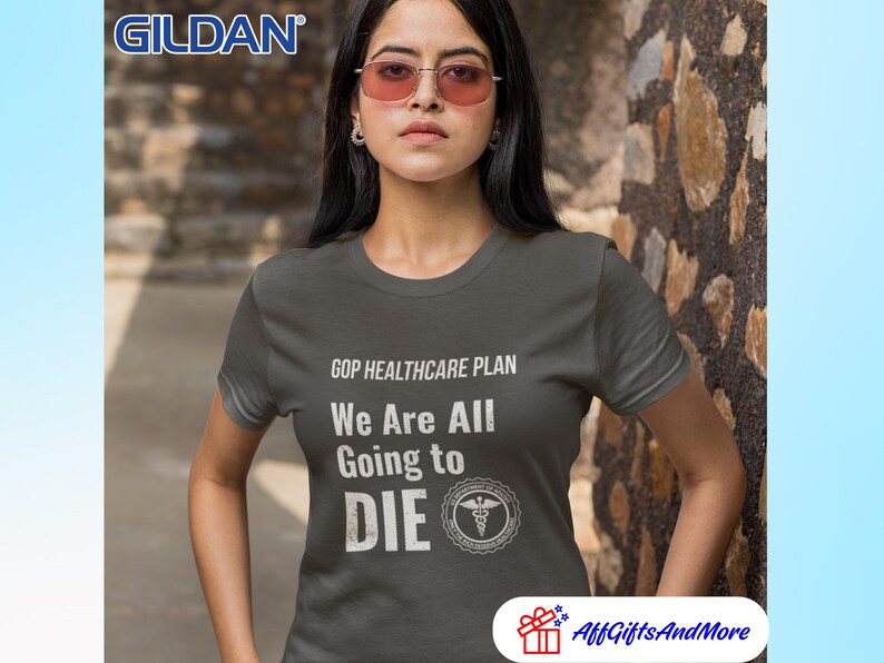 May include: Dark gray t-shirt with the text "GOP HEALTHCARE PLAN We Are All Going to DIE" in white. The shirt features a medical symbol and the Gildan logo. The model is wearing pink-tinted sunglasses.