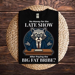 May include: Black t-shirt with a raccoon graphic wearing glasses and a suit behind a desk. Text reads "No Money for the LATE SHOW" and "After Paying the BIG FAT BRIBE?" on the front. The shirt is displayed on a woven mat.