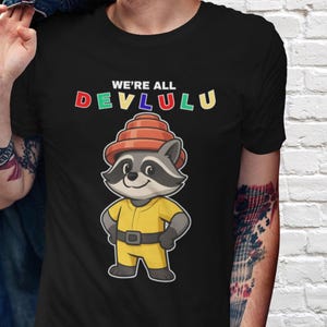 May include: Black t-shirt with the text "WE'RE ALL DEV LULU" in colorful letters. The shirt features a cartoon raccoon wearing a yellow jumpsuit and an orange hat. The raccoon has its hands on its hips.