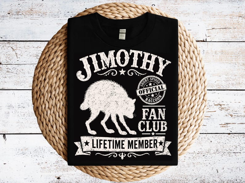 May include: A black t-shirt featuring a white graphic of a raccoon and the text JIMOTHY OFFICIAL SHORT SPINED RACCOON FAN CLUB LIFETIME MEMBER. The design includes a circular badge, stars, and decorative flourishes surrounding the central animal illustration.