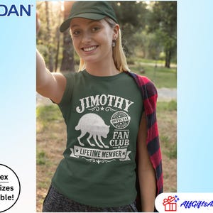 May include: A dark green short-sleeved t-shirt featuring a white graphic of a raccoon and the text JIMOTHY OFFICIAL RACCOON FAN CLUB LIFETIME MEMBER. The shirt is worn with a red and black plaid flannel shirt draped over one shoulder.