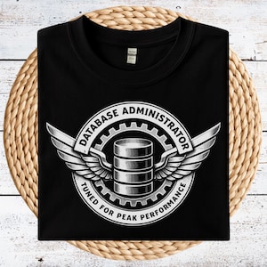 May include: Black t-shirt featuring a white and silver graphic. The graphic includes a database server illustration, wings, and the text "DATABASE ADMINISTRATOR" and "TUNED FOR PEAK PERFORMANCE". The shirt is folded on a woven surface.