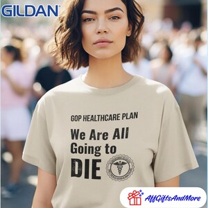 May include: A beige Gildan t-shirt with the text "GOP HEALTHCARE PLAN We Are All Going to DIE" in black. The shirt also features a medical symbol. The image is of a person wearing the shirt.