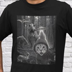 May include: Black t-shirt featuring a grayscale graphic of a cat in a hoodie, a T-Rex, a Bigfoot, a UFO, and a pterodactyl in a forest setting. The cat is the focal point, with the other elements in the background.