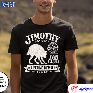 May include: A black short-sleeved t-shirt featuring a white graphic of a raccoon. The text reads JIMOTHY, OFFICIAL SHORT-SPINED RACCOON FAN CLUB, and LIFETIME MEMBER. The design includes a circular stamp and a banner element on the front chest.