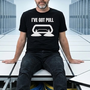 May include: Black t-shirt with the text "I'VE GOT PULL" and a white graphic of a suction cup tool. The person is wearing black jeans and sitting on a white tiled floor. The background has rows of white panels.