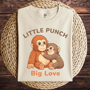 May include: Cream-colored t-shirt featuring a cartoon illustration of two monkeys hugging. The text "LITTLE PUNCH" is arched above the monkeys, with "Big Love" printed below in orange. The shirt is displayed on a woven mat.